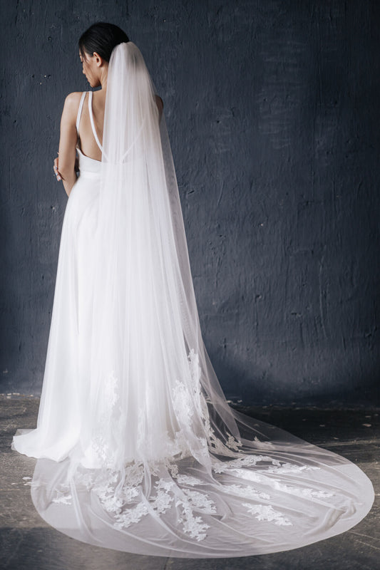 Bride wearing a long, elegant white wedding dress with a matching veil against a dark background.