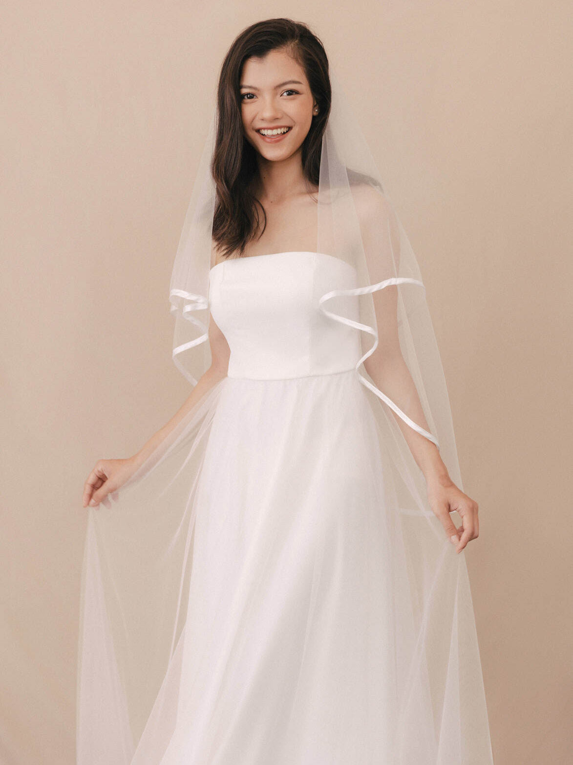 Woman wearing a strapless white wedding dress with a sheer veil against a beige background