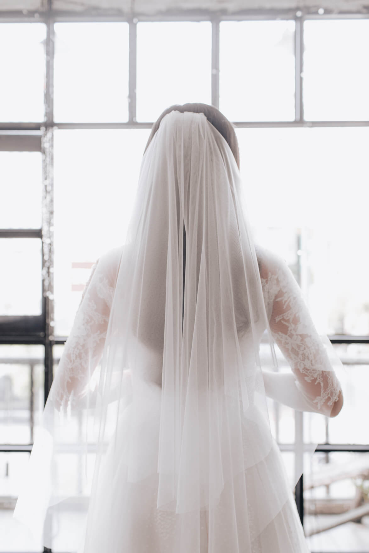 Person wearing a long white wedding dress with a veil in front of a window.