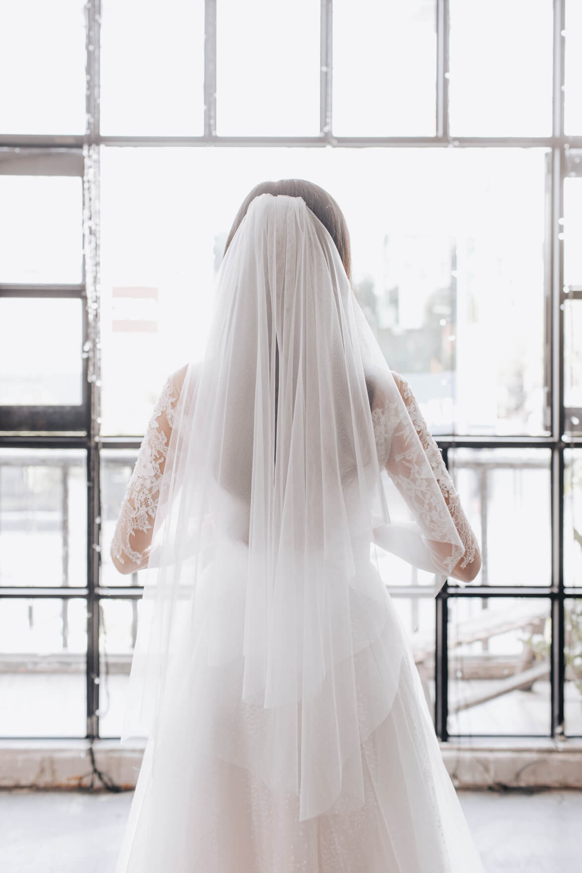 Person wearing a long white wedding dress with a veil in front of a glass wall.