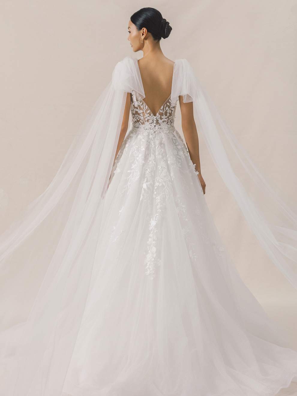 Back view of a woman wearing a white wedding dress with a sheer cape against a beige background