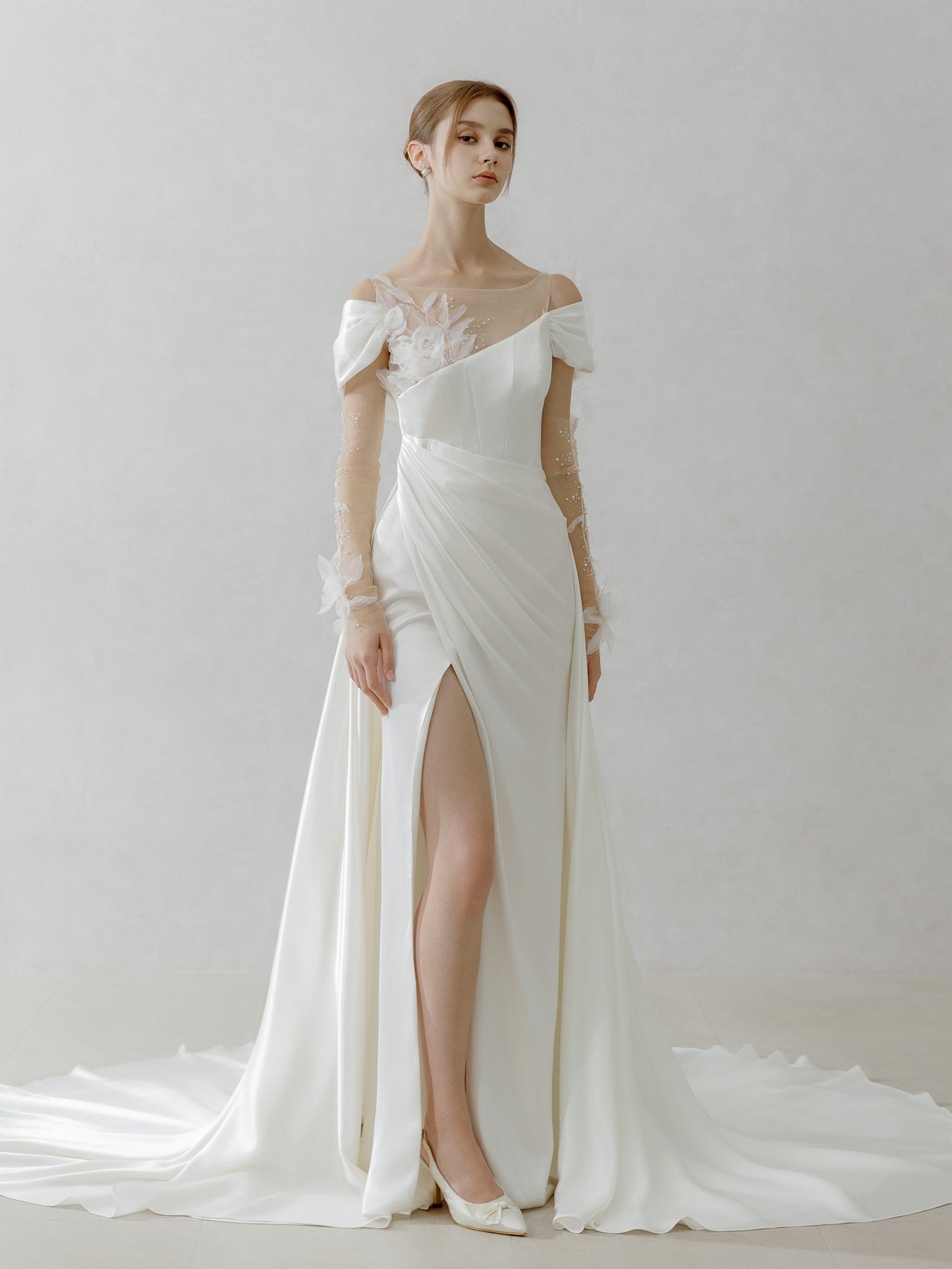 Woman wearing a white off-shoulder wedding dress with a high slit on a plain background