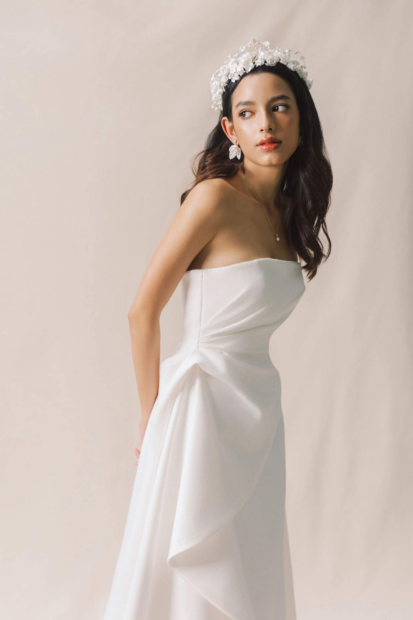 Woman in a strapless white wedding dress with a floral headpiece against a beige background