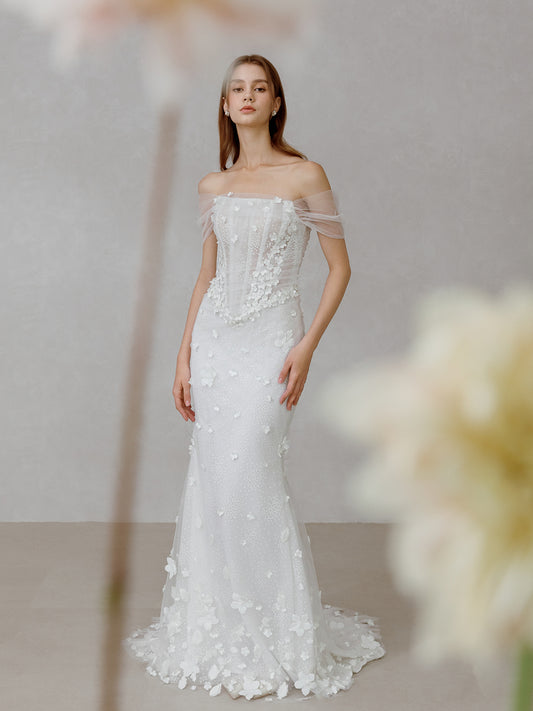 Woman wearing a white off-shoulder wedding dress with floral details against a neutral background