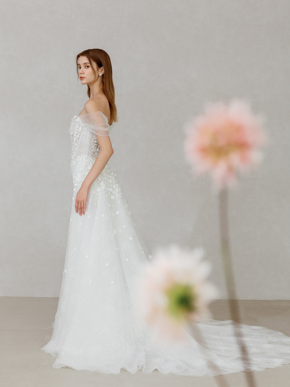 Woman in a white wedding dress standing against a neutral background with flowers in the foreground.