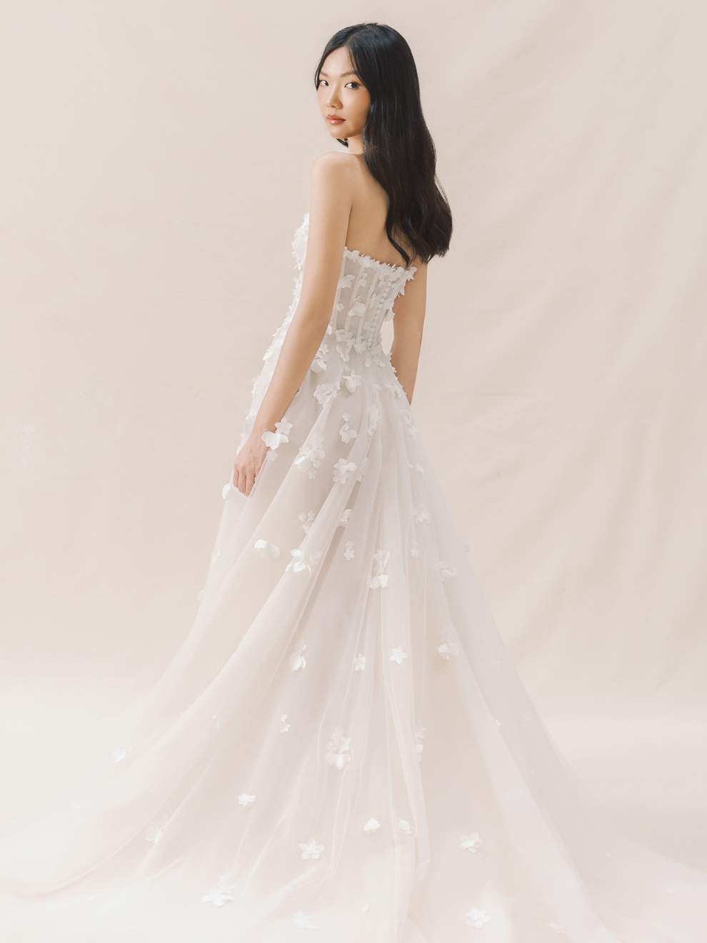 Woman wearing a strapless white wedding dress with floral details against a light pink background