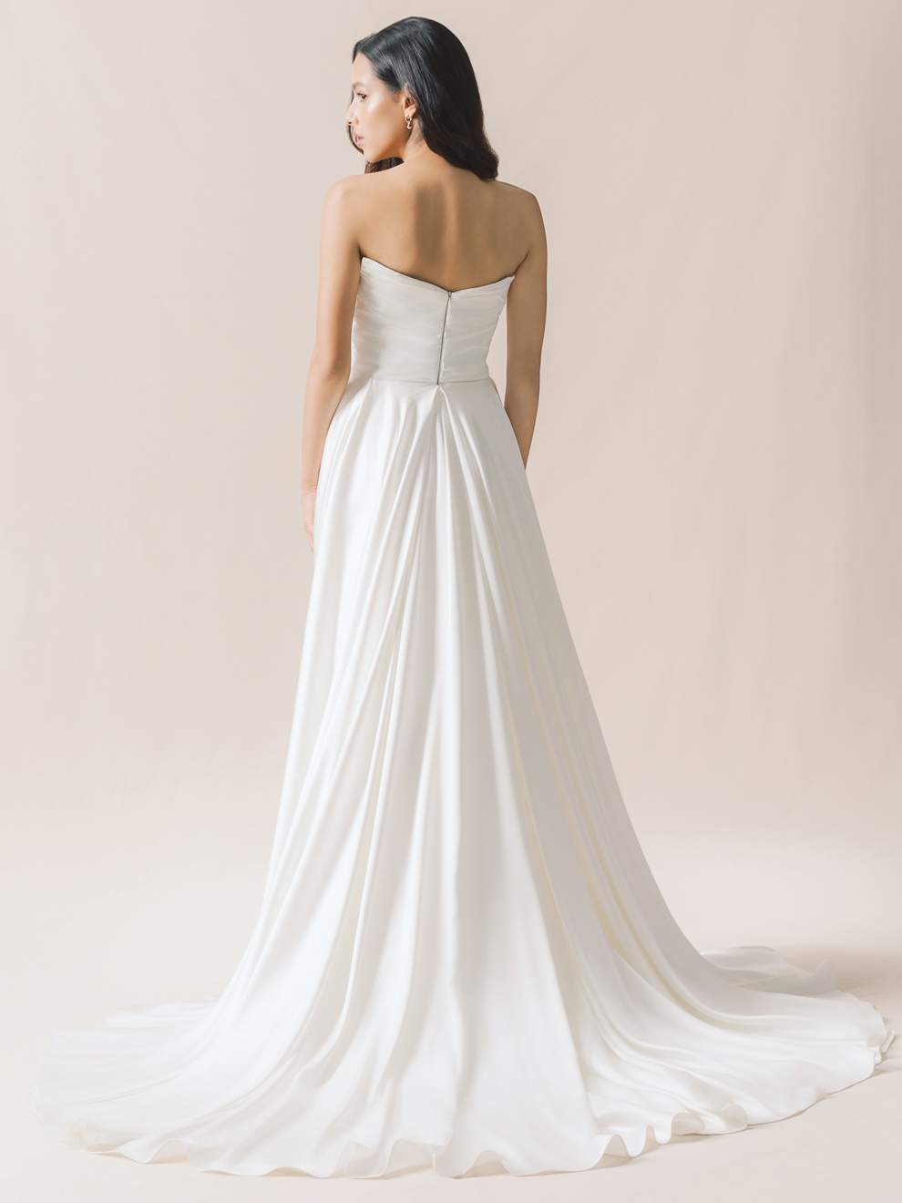 Woman wearing a strapless white wedding dress with a long train on a plain background