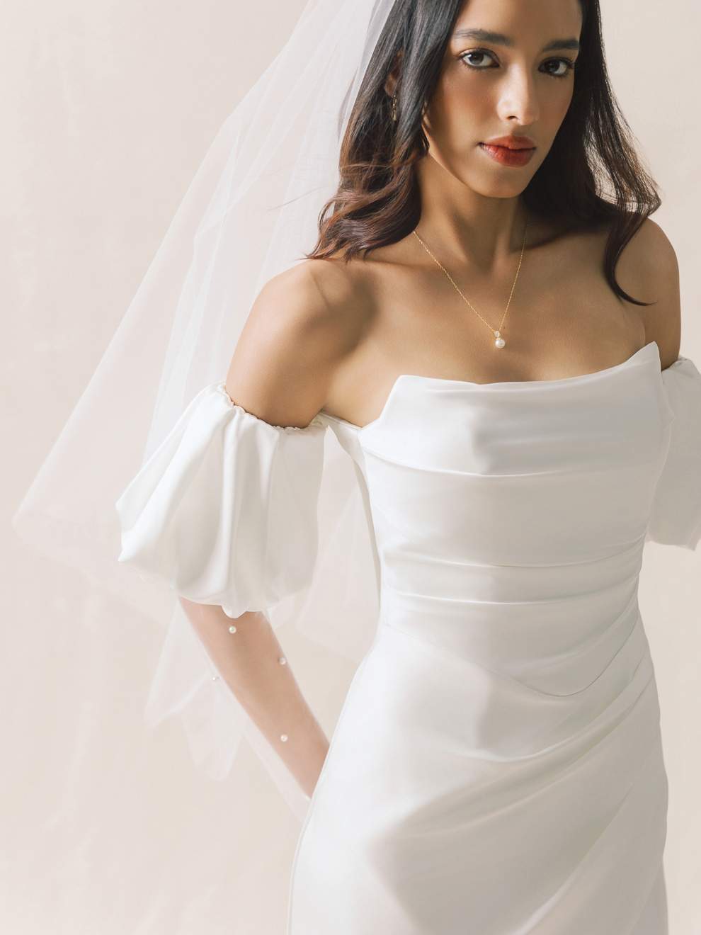 Woman wearing a white off-shoulder dress with a veil against a plain background
