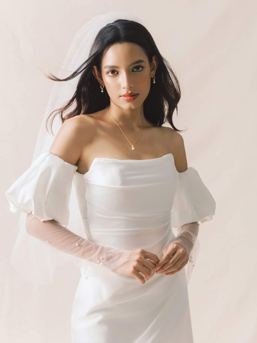 Woman wearing a white wedding dress with sheer sleeves against a plain background