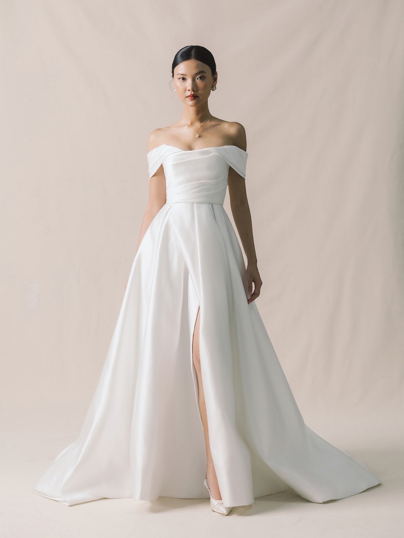 Woman wearing a white off-shoulder gown with a high slit on a plain background