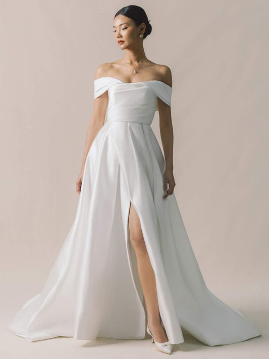 Woman wearing a white off-shoulder gown with a high slit on a plain background