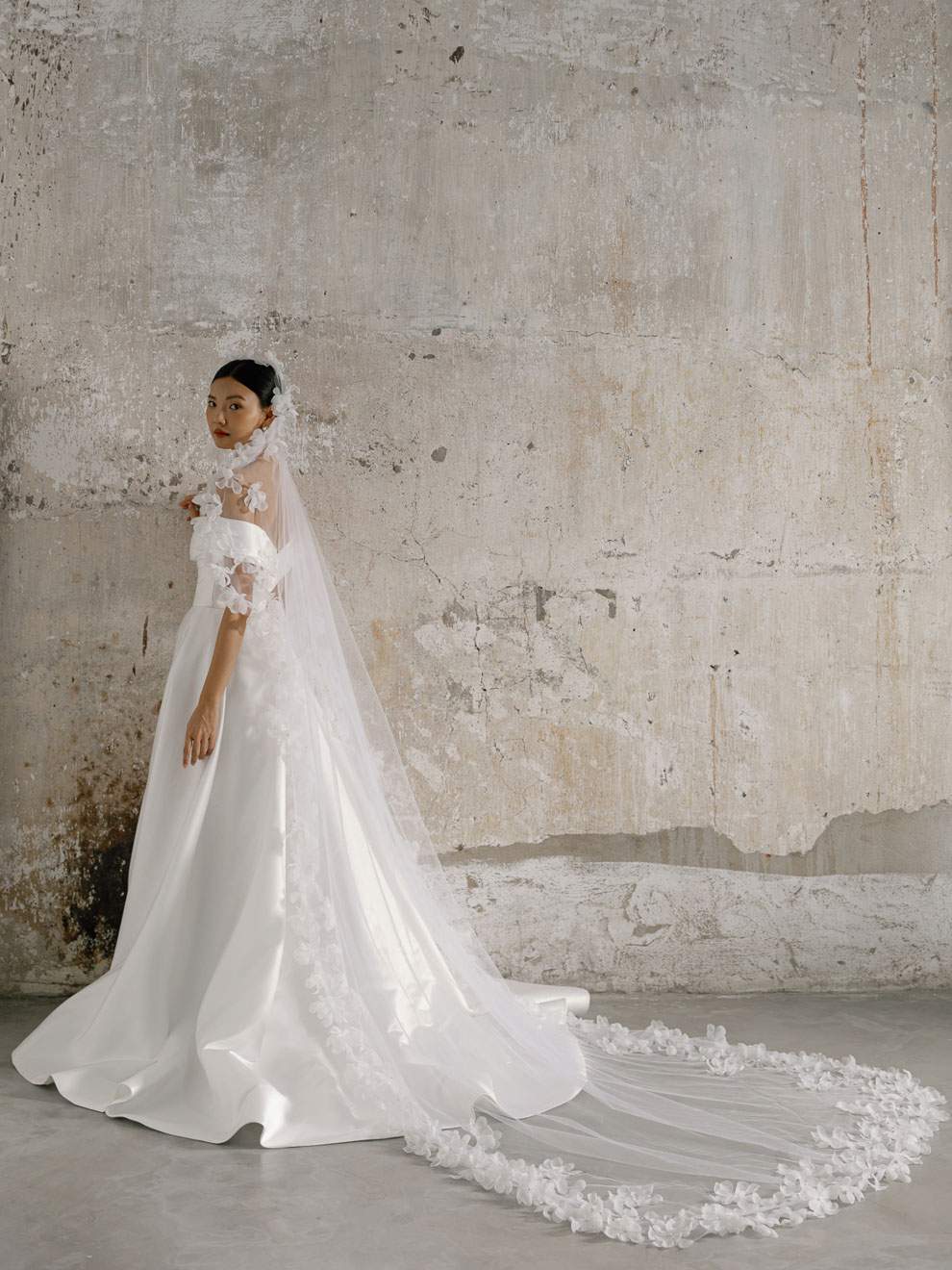 Woman in a white wedding dress with a long veil against a textured wall.