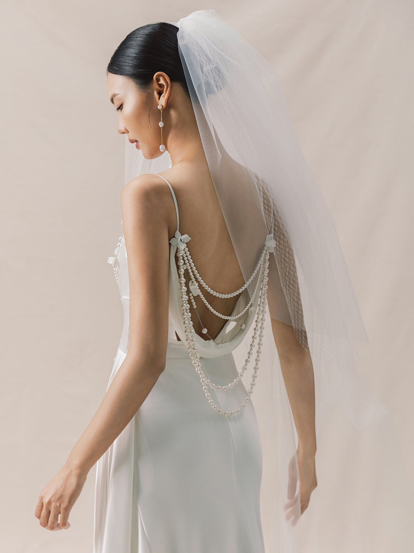 Bride wearing a white wedding dress with intricate detailing and a veil against a neutral background