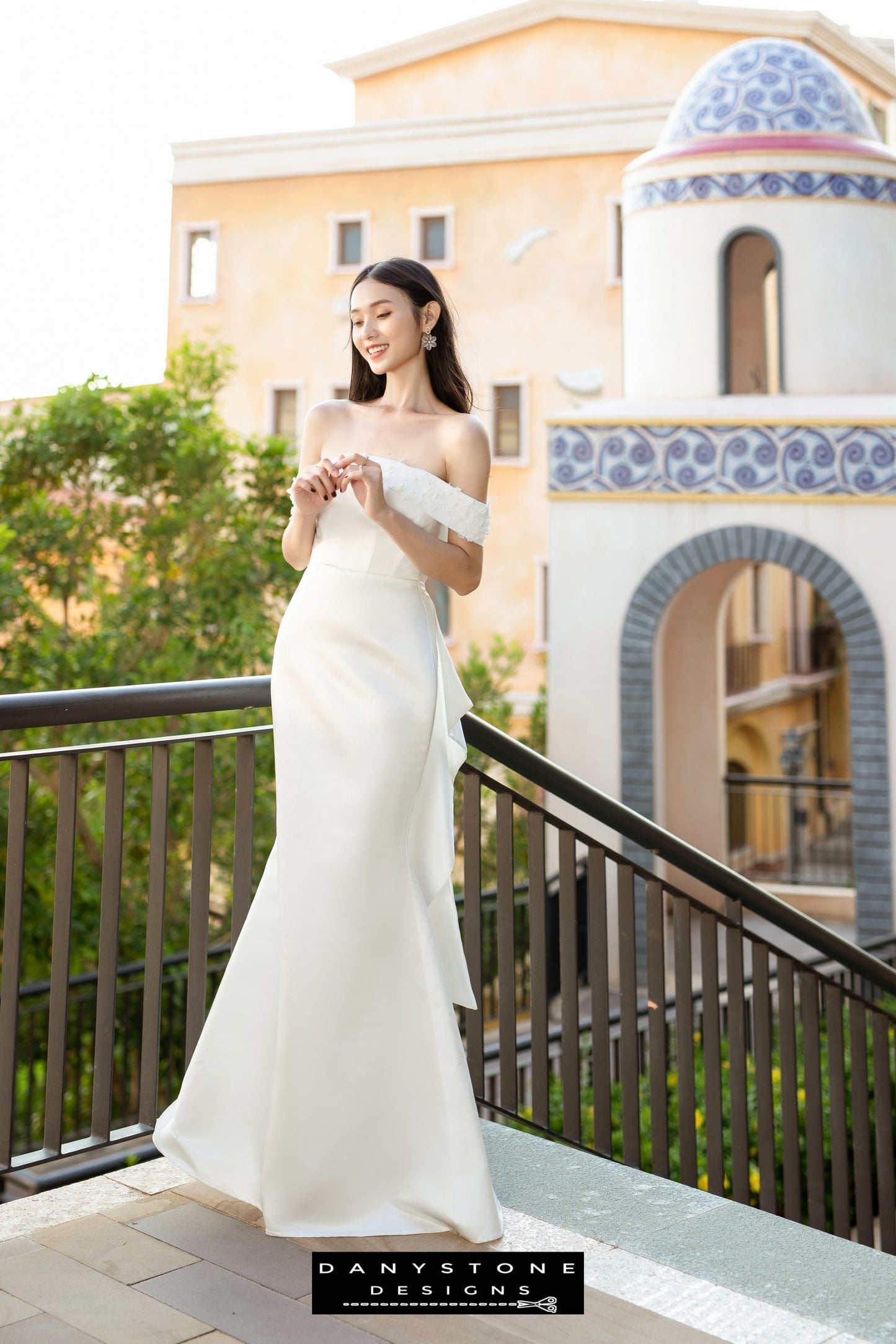 Bride in a flat shoulder fishtail wedding dress with 3D floral accents, smiling and looking elegant on a balcony.