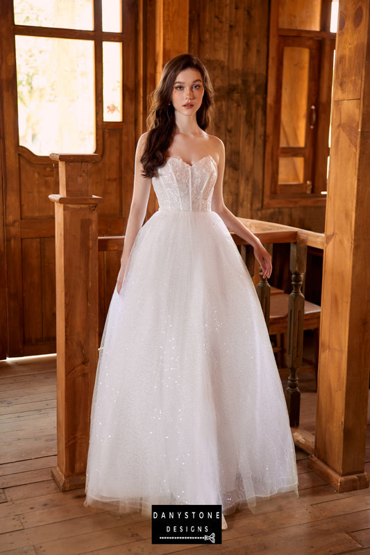 Bride wearing an ethereal strapless wedding dress with lace bodice and mesh chiffon overlay, standing by a wooden railing.