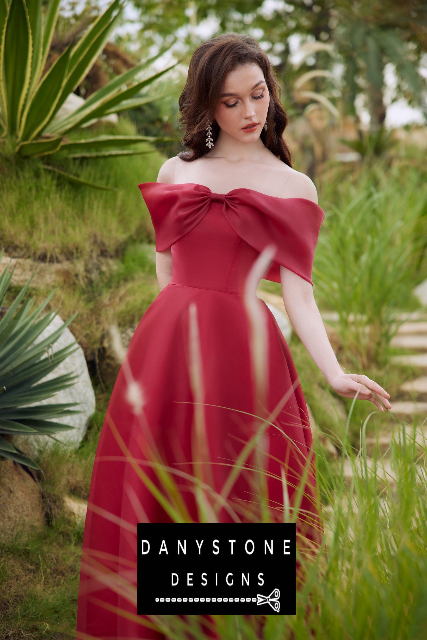 Model showcasing a red off-shoulder silk chiffon dress with a bow in a natural setting