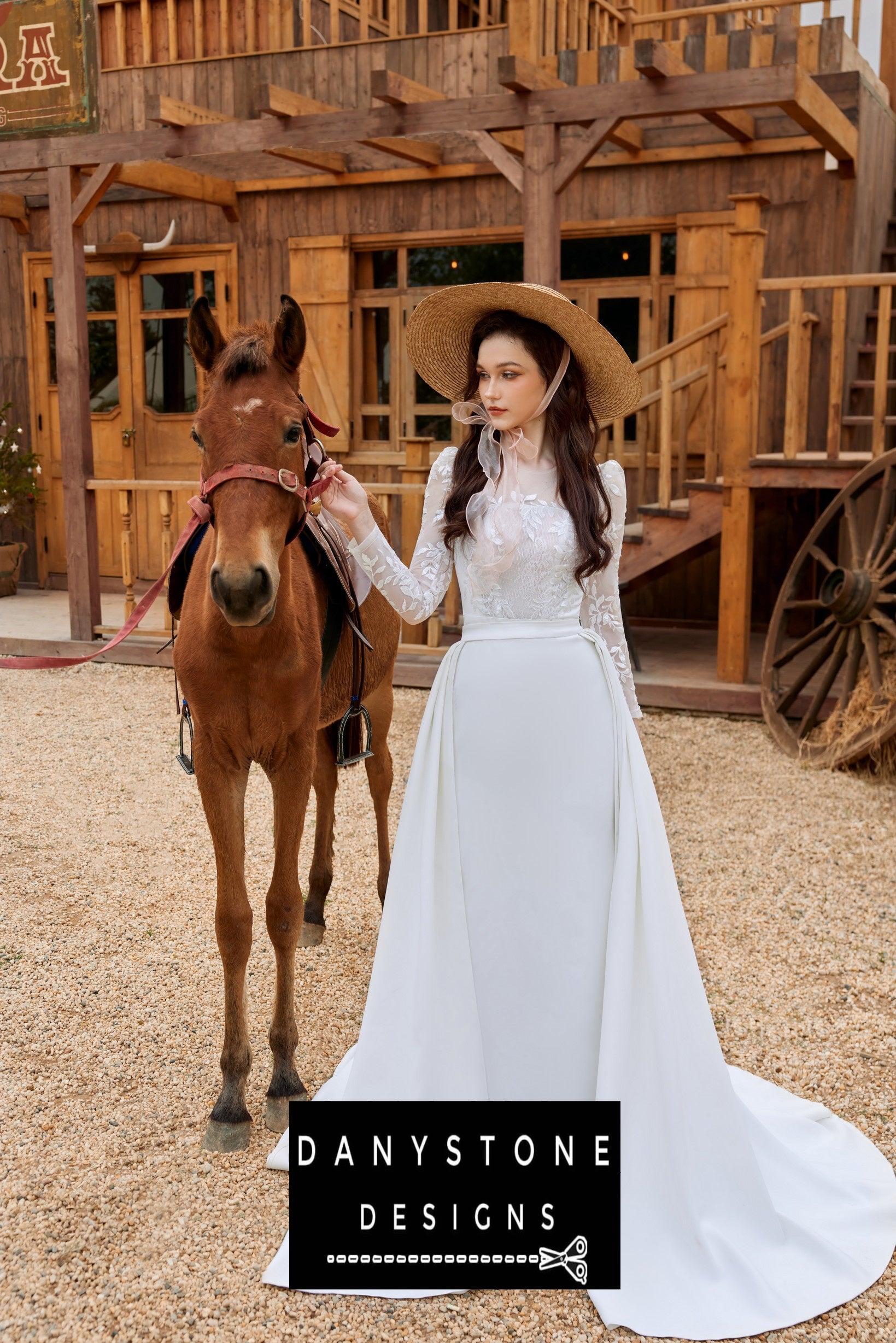 Bride holding a horse, wearing a long-sleeved fishtail wedding dress with leaf motifs and a wide-brimmed hat, standing in front of a rustic building.