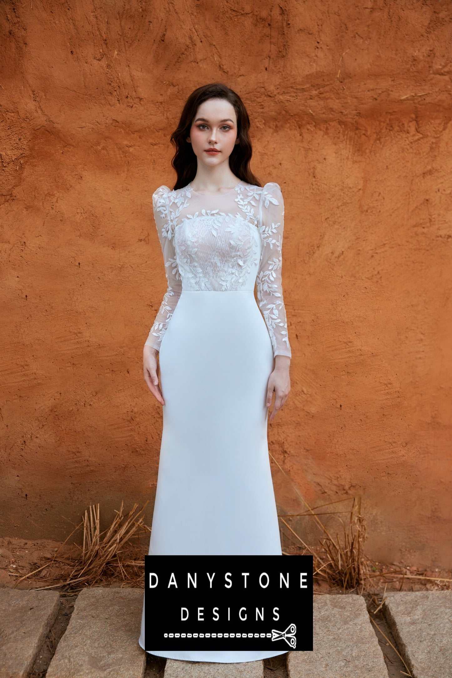 Bride standing against an orange wall in a long-sleeved fishtail wedding dress with leaf motifs and 3D flowers, looking directly at the camera.