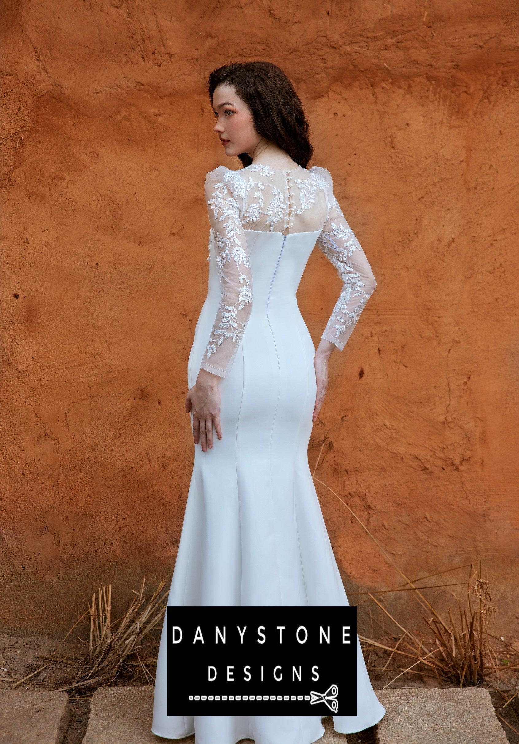 Back view of the bride in a long-sleeved fishtail wedding dress with leaf motifs and 3D flowers, standing against an orange wall.