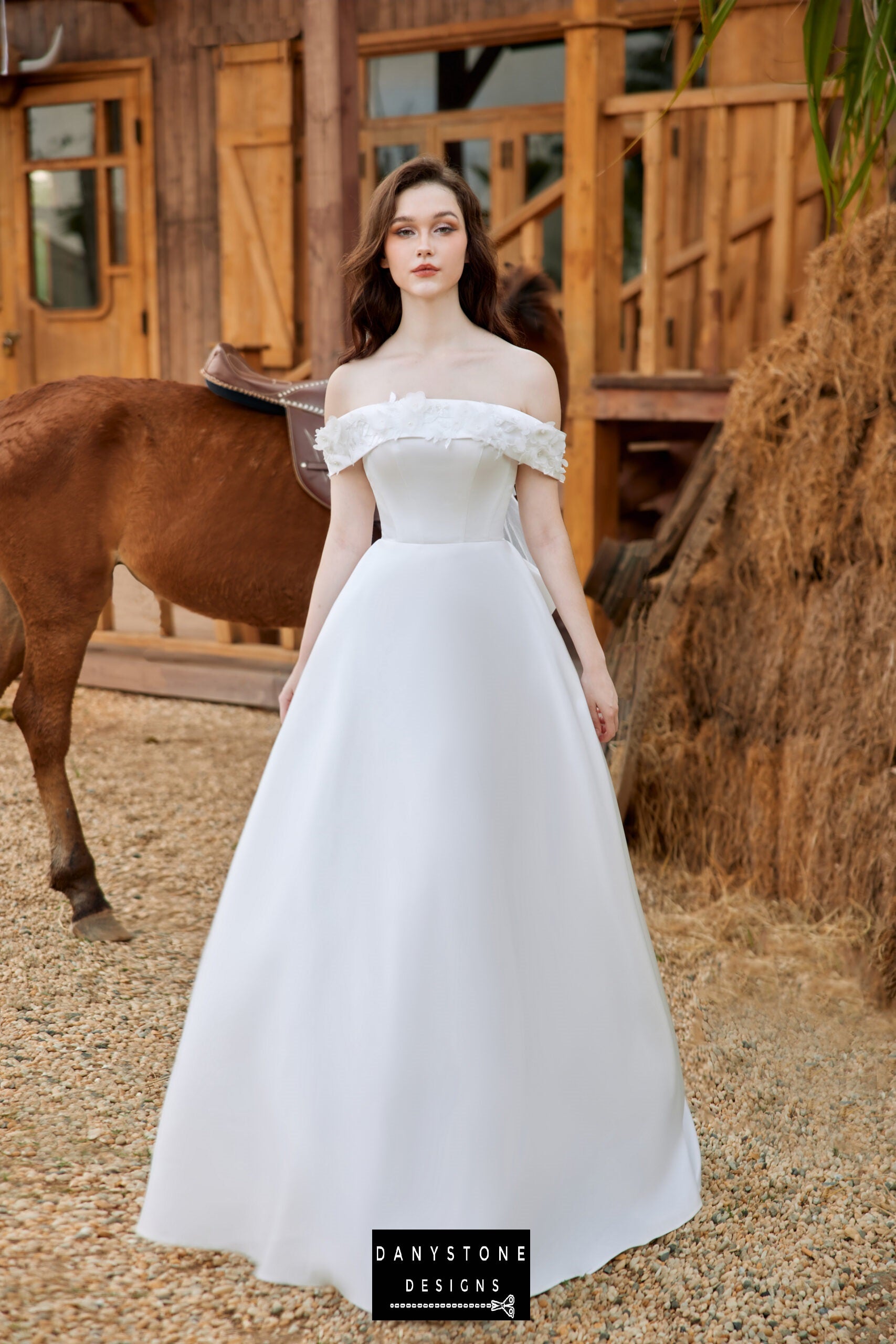 Elegant off-the-shoulder satin wedding dress with floral accents by Danystone Designs - front view with horse in the background.