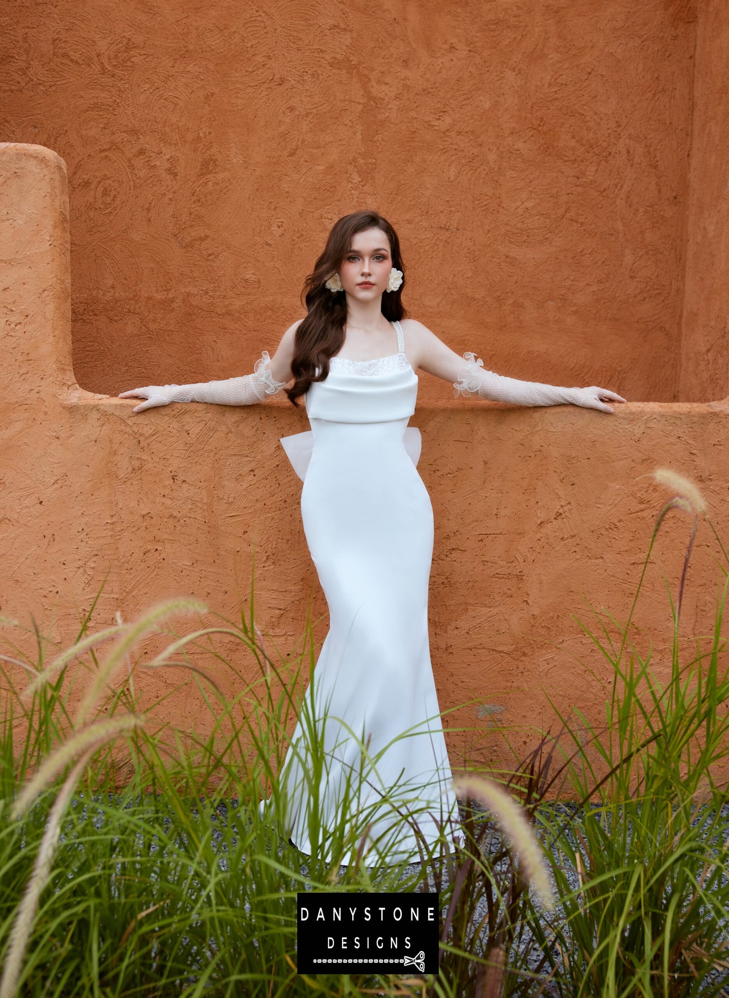 Bride posing in a garden wearing a satin neckline wedding dress with pearl strap back, with her arms resting on an orange wall.