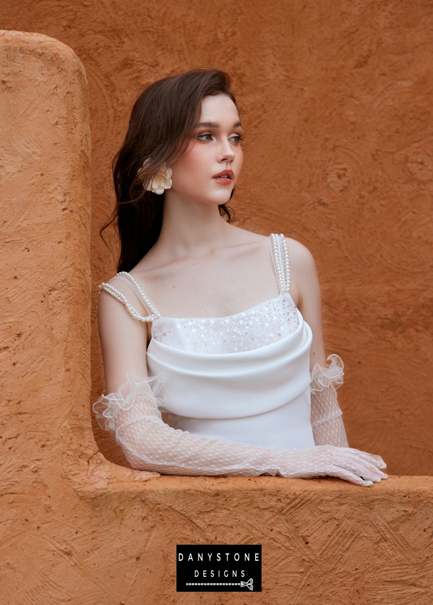 Bride peeking over an orange wall in a satin neckline wedding dress with pearl strap back, looking into the distance