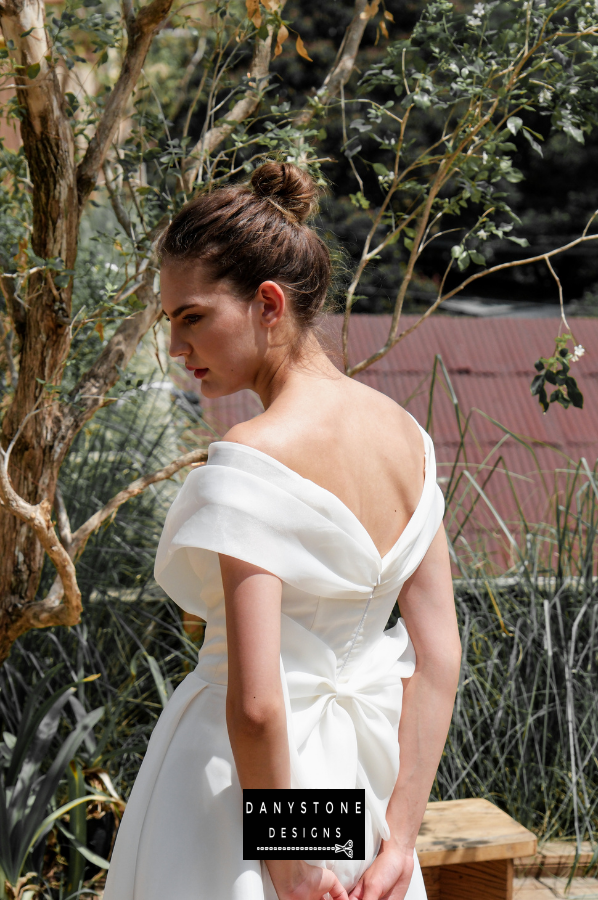 Chic Off-the-Shoulder Bridal Gown with Large Bow - Model standing in a picturesque outdoor setting, showcasing the unique bow detail and the sophisticated design of the gown.