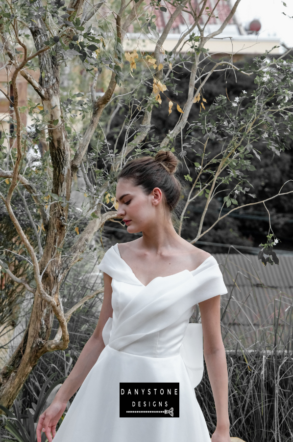 Classic Off-the-Shoulder Bridal Dress with Bow Detail - Model presenting the side view of the gown, showcasing the elegant pleats and captivating bow at the back.