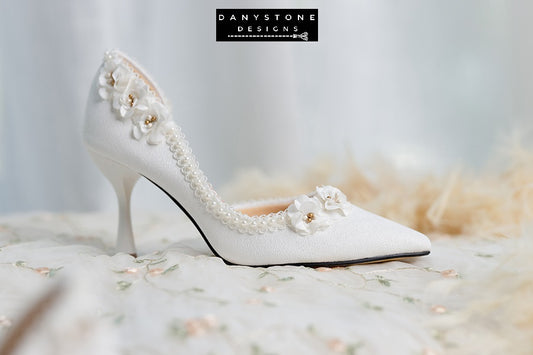 Elegant wedding shoes with open waist, pearl trim, and floral accents, displayed on a white fur background.