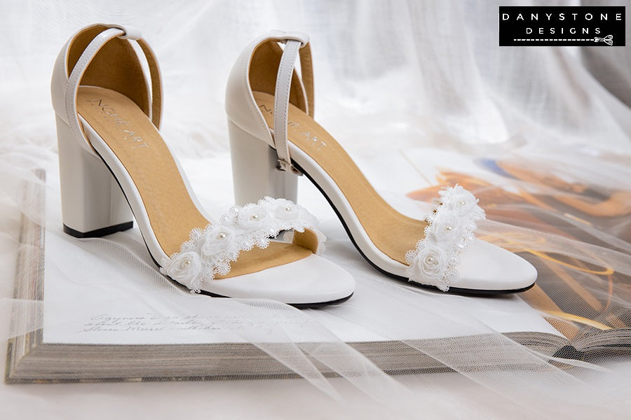 Matte white leather heels with white rose lace trim, displayed on an open book with a veil.