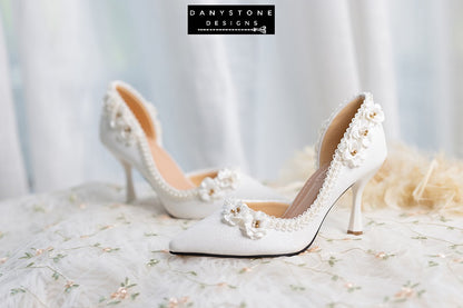 Front view of wedding shoes with open waist, pearl trim, and floral accents, displayed on a floral fabric.