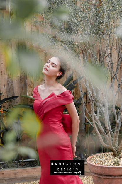 Fashion model in a vibrant red silk gown with a sculpted pleat design, outdoors among wooden and plant decor.