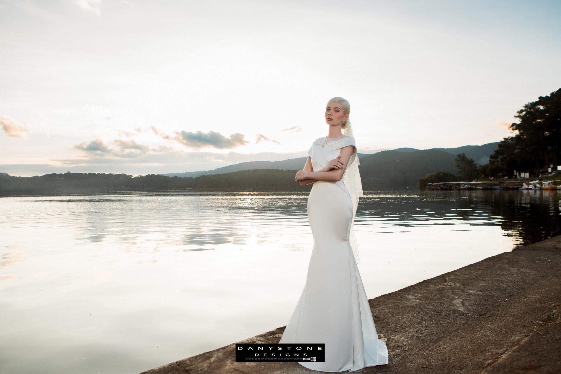 Image 3: Full-length view of a bride in a satin mermaid wedding dress with floral appliqué, against a scenic lake background.