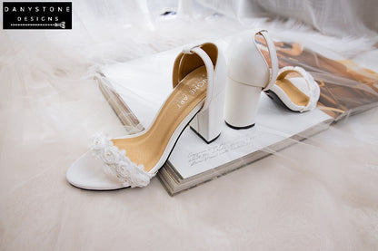 Matte white leather heels with white rose lace trim, displayed on an open book with a veil.