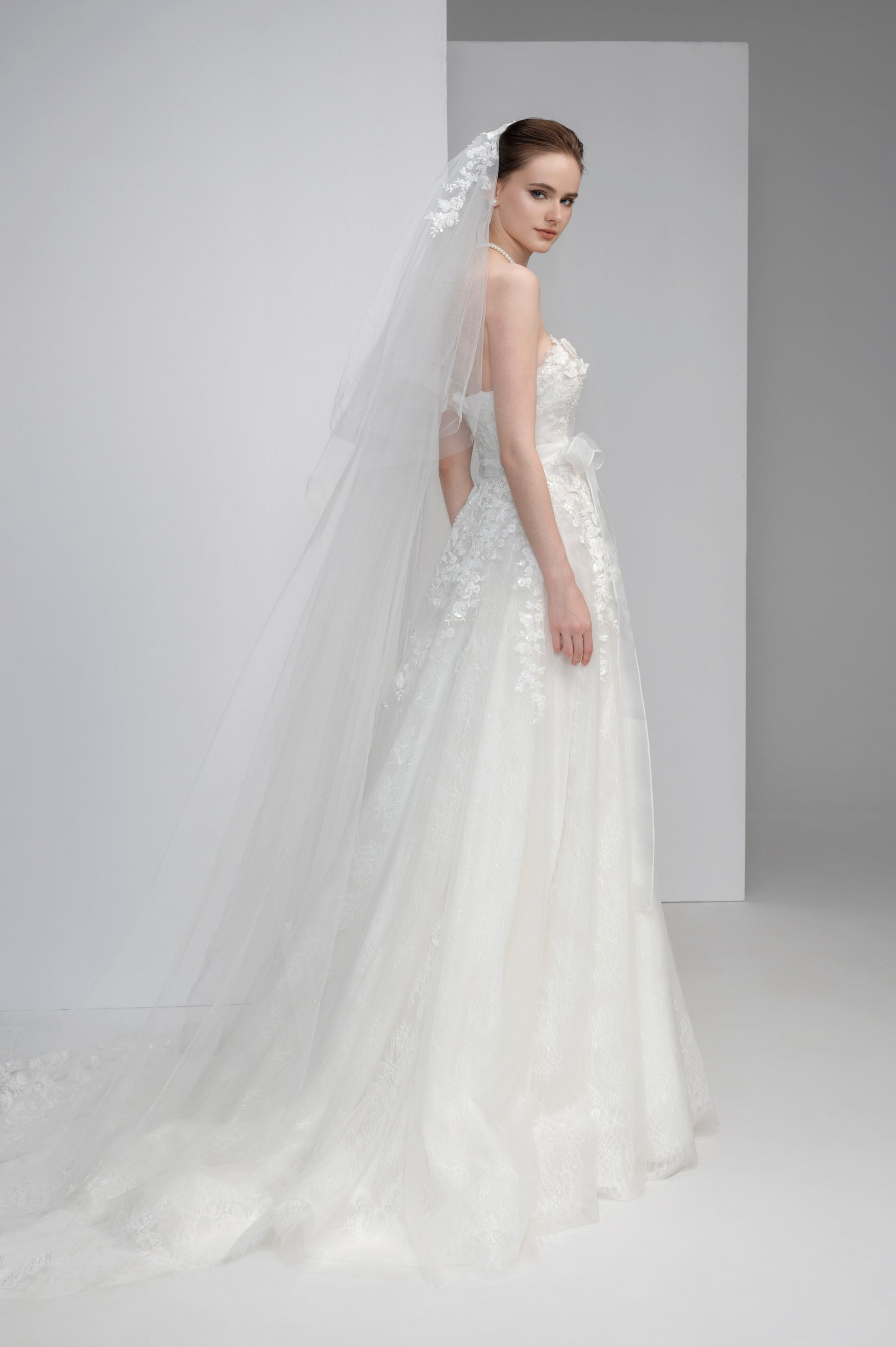 Model wearing a white wedding dress with a long veil against a plain background