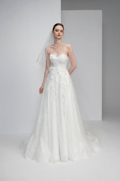 Model wearing a strapless white wedding dress with floral details against a plain background