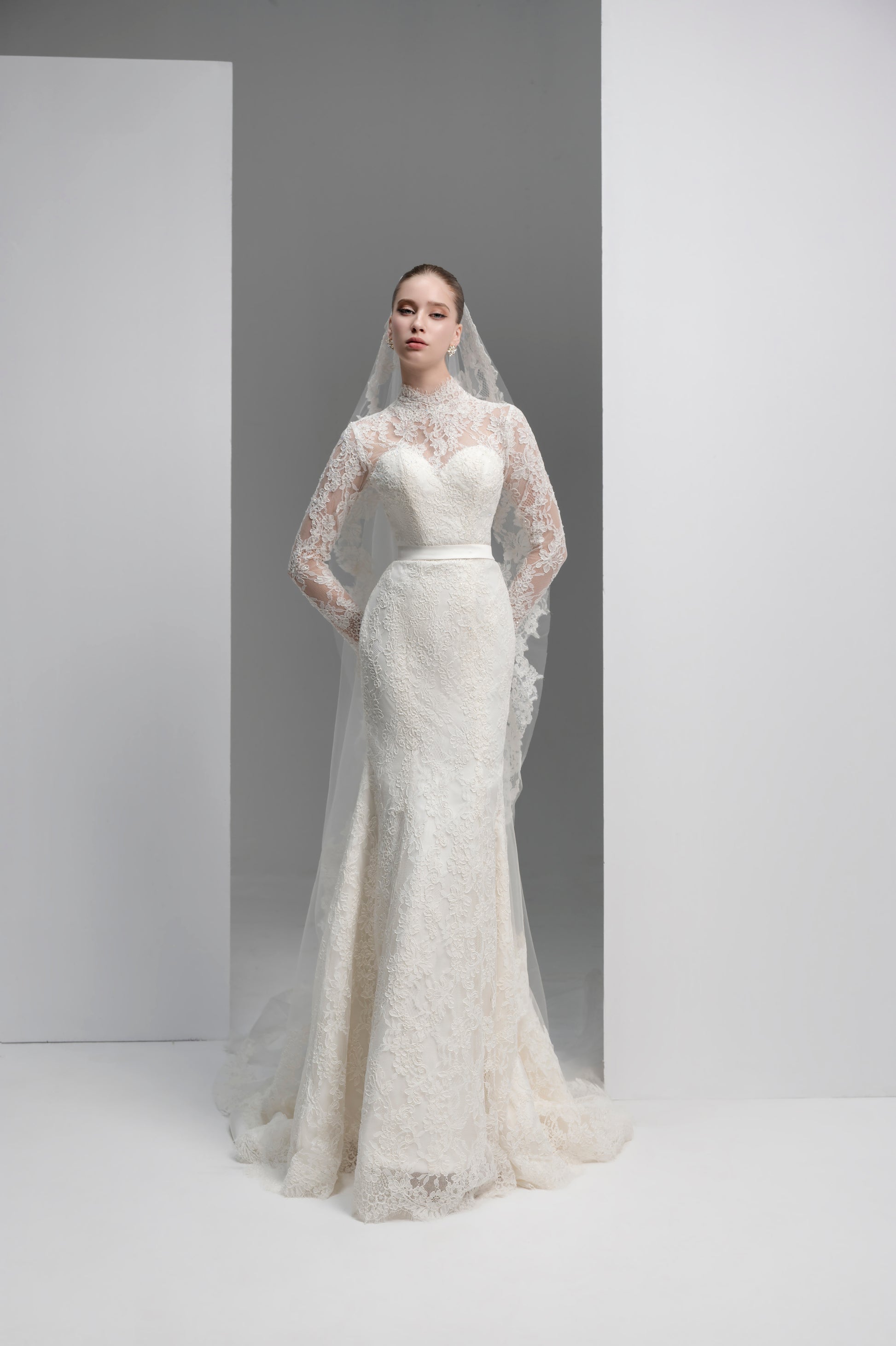 Woman wearing a white lace wedding dress with a long veil against a plain background