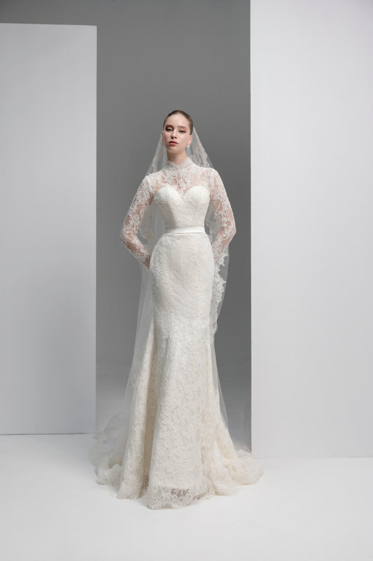 Woman wearing a white lace wedding dress with a long veil against a plain background