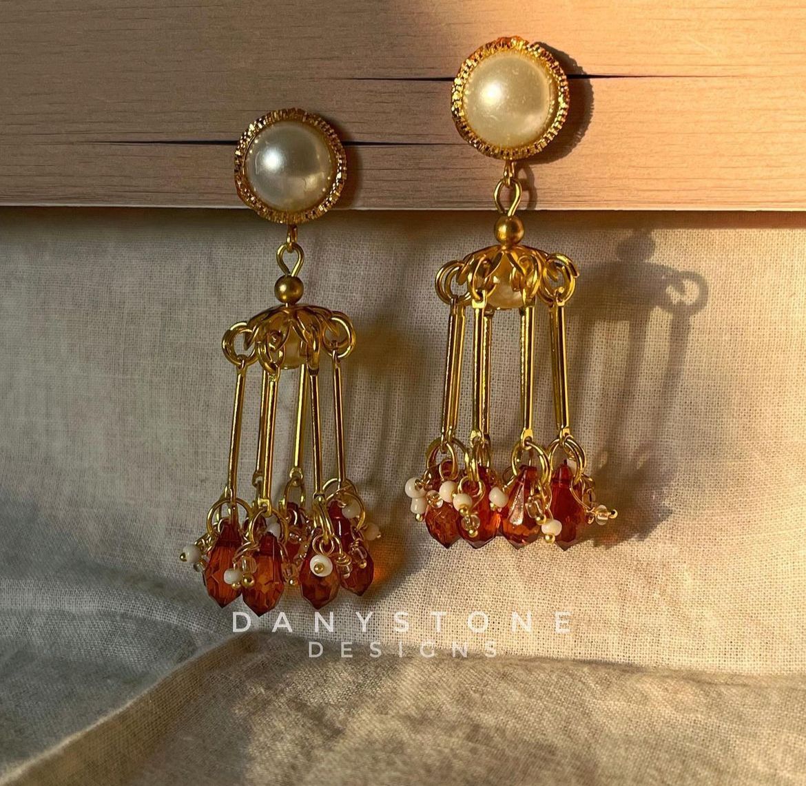 Handmade tassel jhumka earrings with pearl stud, featuring gold-toned tassels and amber and pearl beads, displayed on a soft beige fabric background.