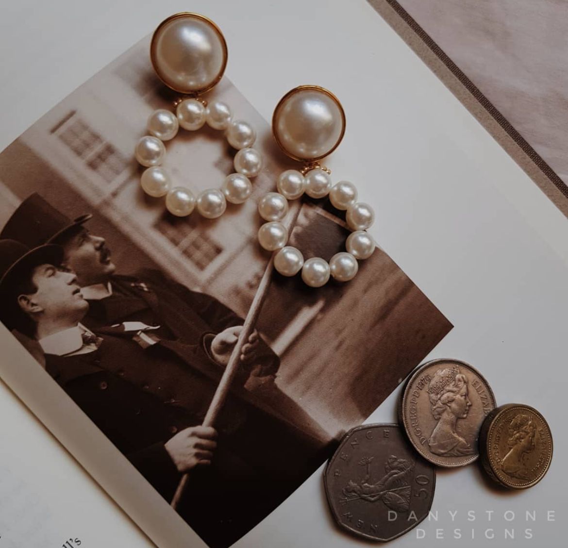 Elegant pearl drop statement earrings with large pearls on a gold-toned base, displayed on an open book with vintage photo and coins.