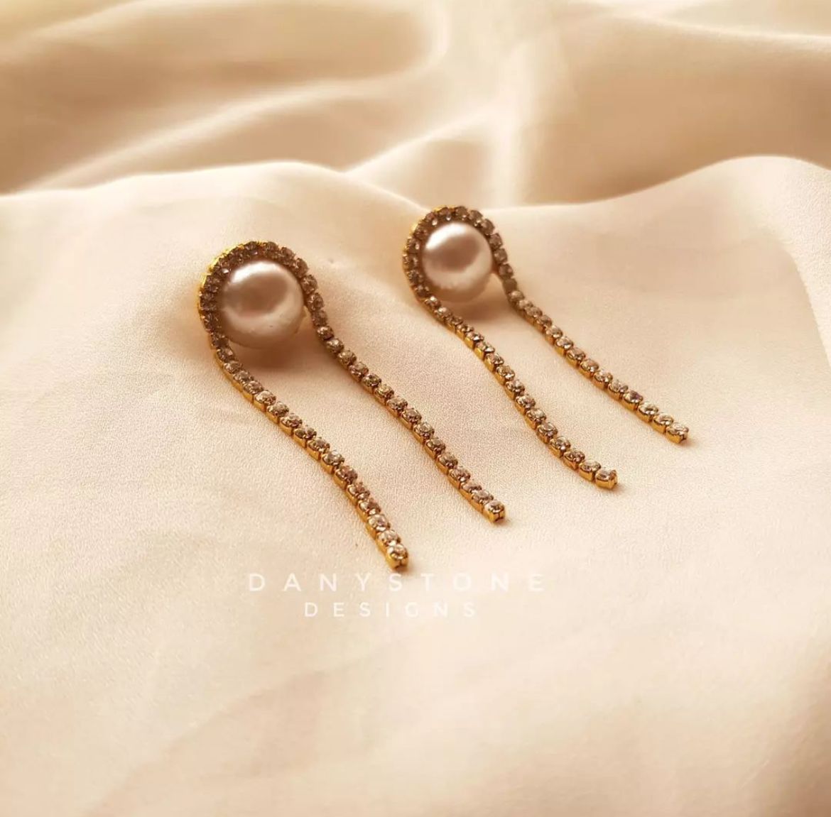 Minimal handcrafted pearl earrings with rhinestones, featuring a delicate pearl and shimmering rhinestone chain, displayed on a soft beige fabric background.