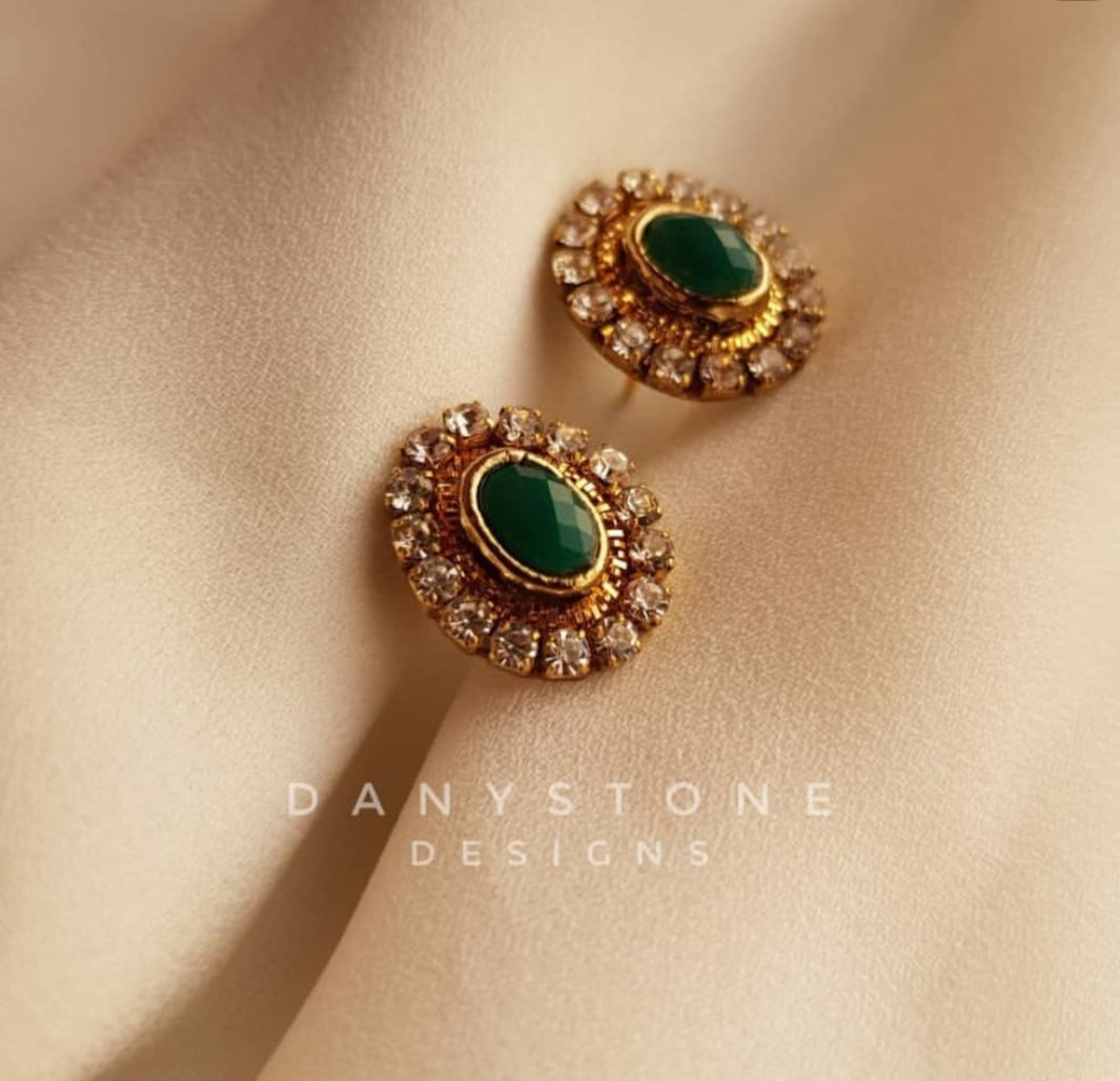 Handcrafted emerald green stud earrings surrounded by rhinestones, displayed on a soft beige fabric background.