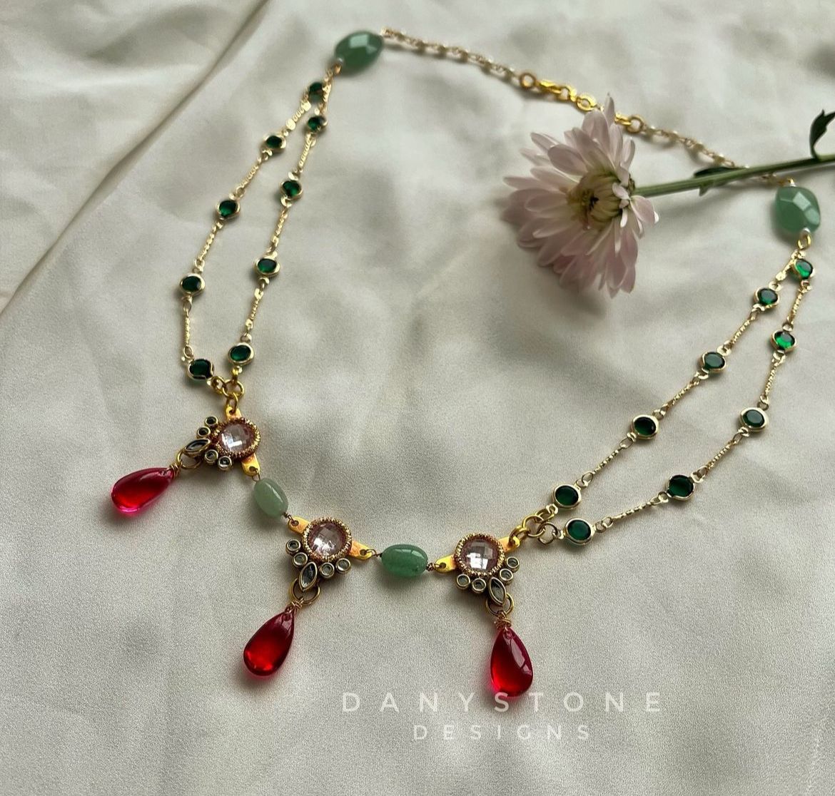 Artisanal Victorian-Indian fusion necklace featuring red drops, green gemstones, and intricate polki-like details, displayed on a soft beige fabric background with a flower.