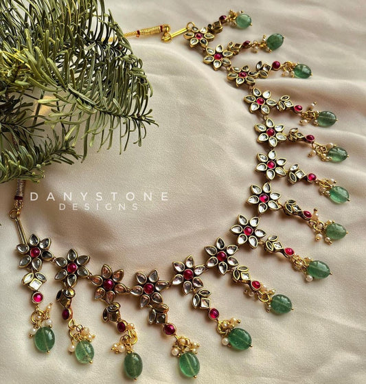 Premium handcrafted polki necklace with bright pink stones and jade drops, displayed on a soft beige fabric background with a sprig of greenery.