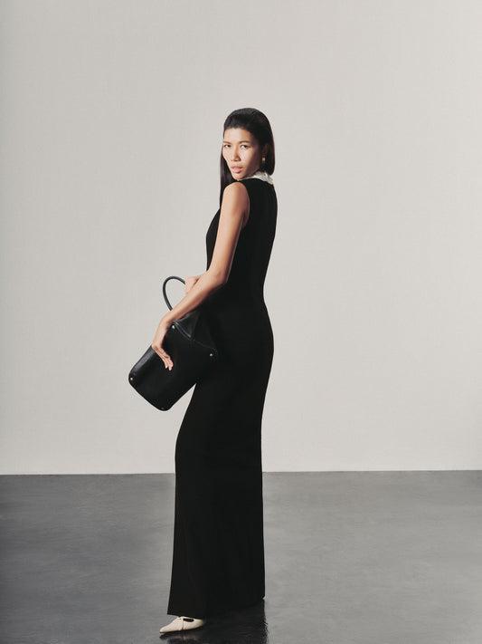 Woman in a black dress holding a black handbag against a plain background