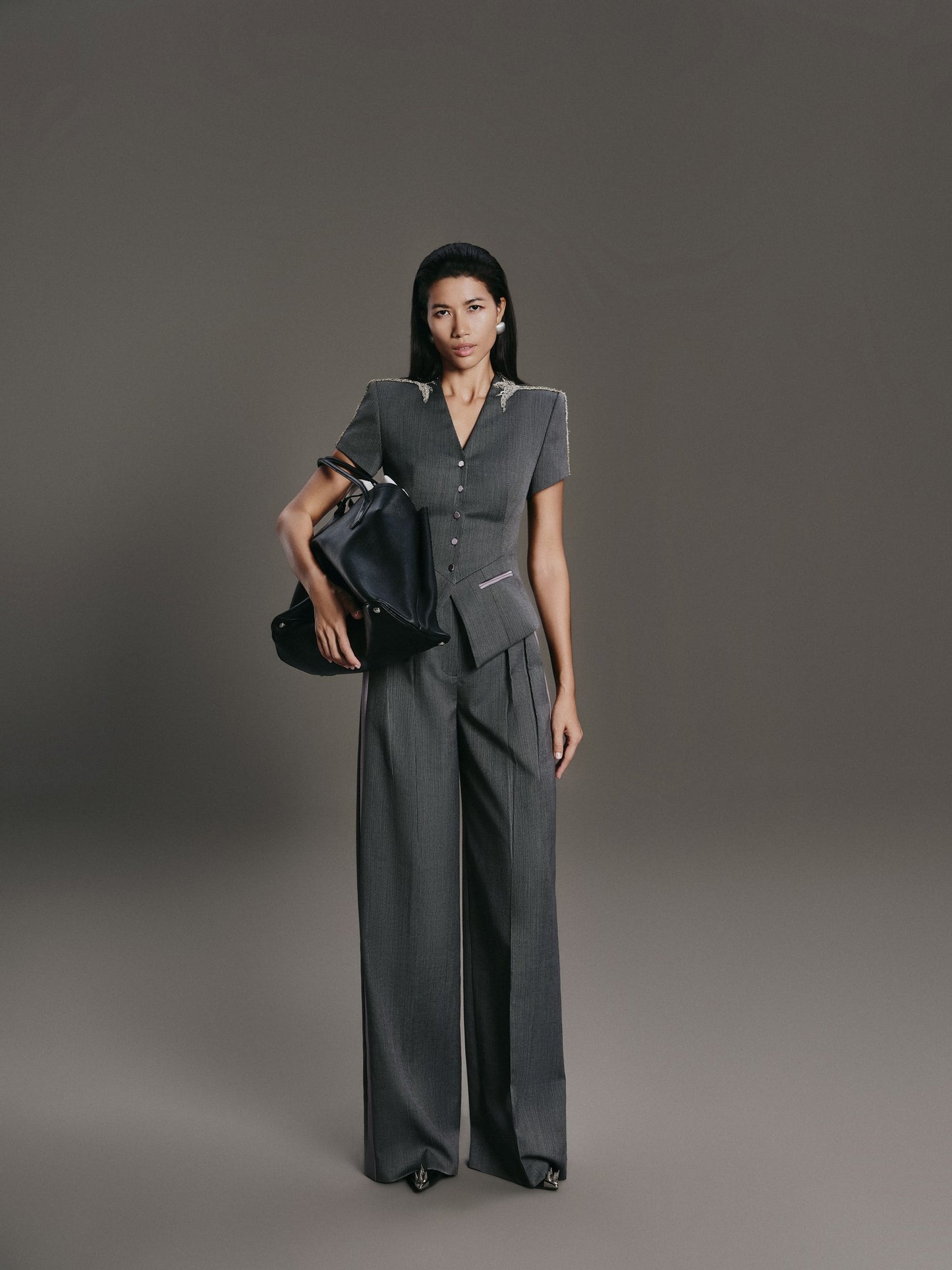 Woman in a gray outfit holding a black handbag against a gray background
