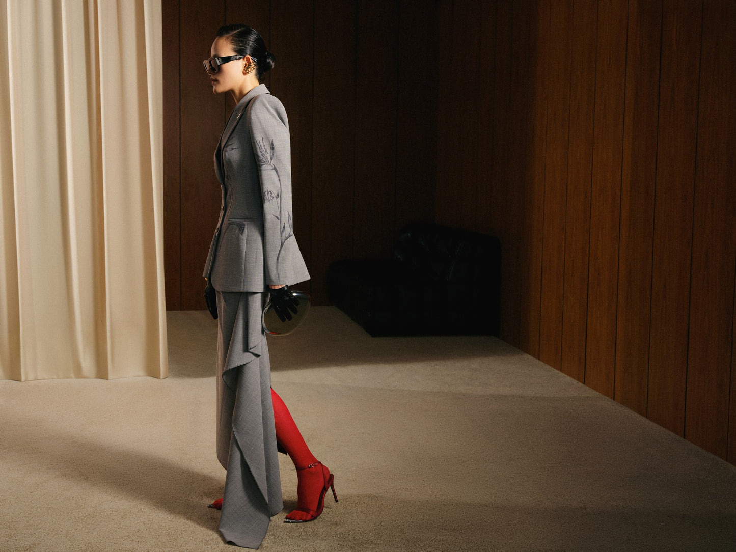Woman in a gray suit and red high heels standing in a room with beige curtains and wooden walls.