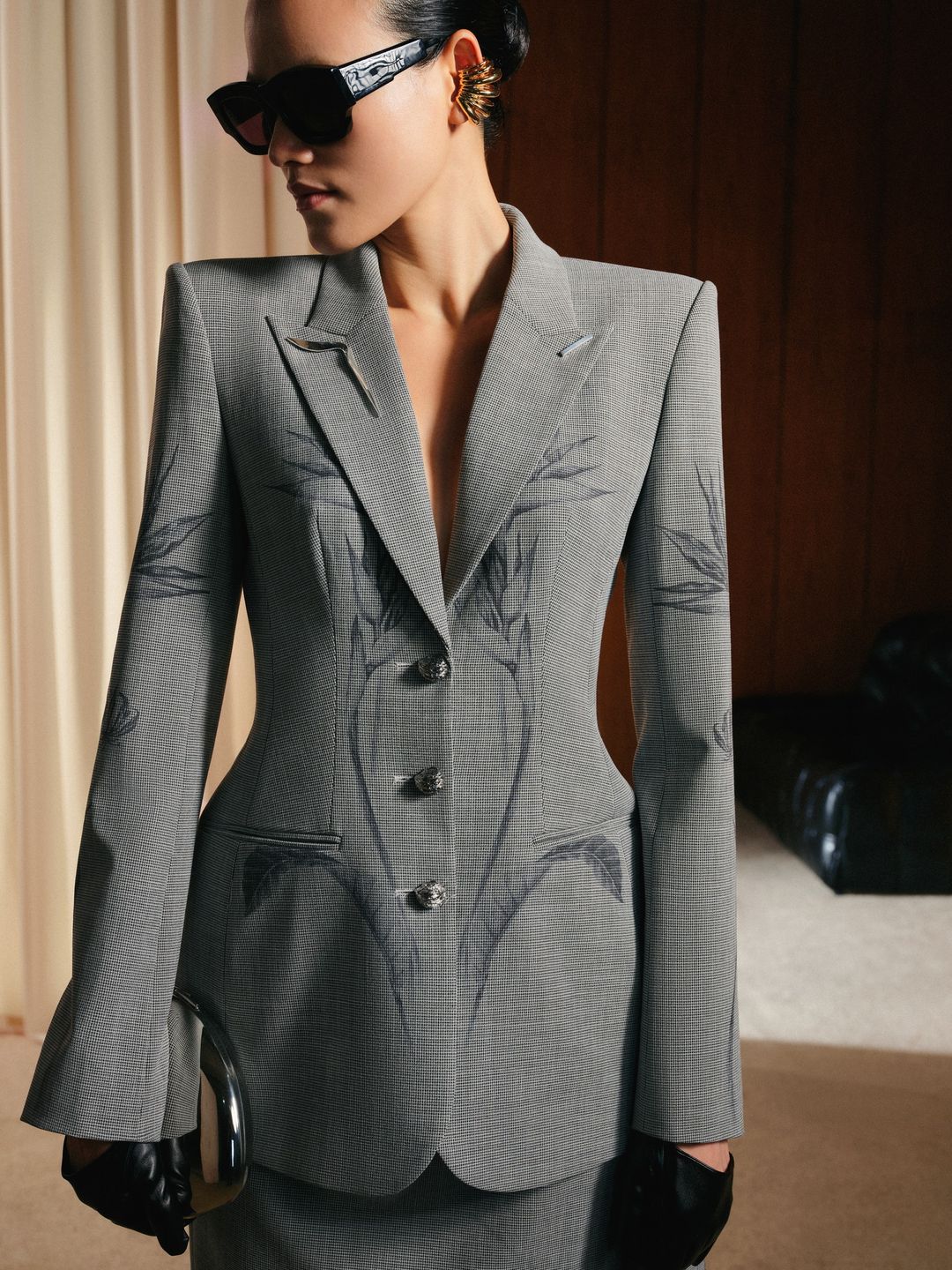 Woman wearing a gray suit with sunglasses and gloves indoors.