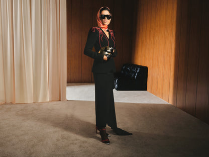 Person in a black outfit with a red scarf standing in a room with beige curtains and wooden walls.