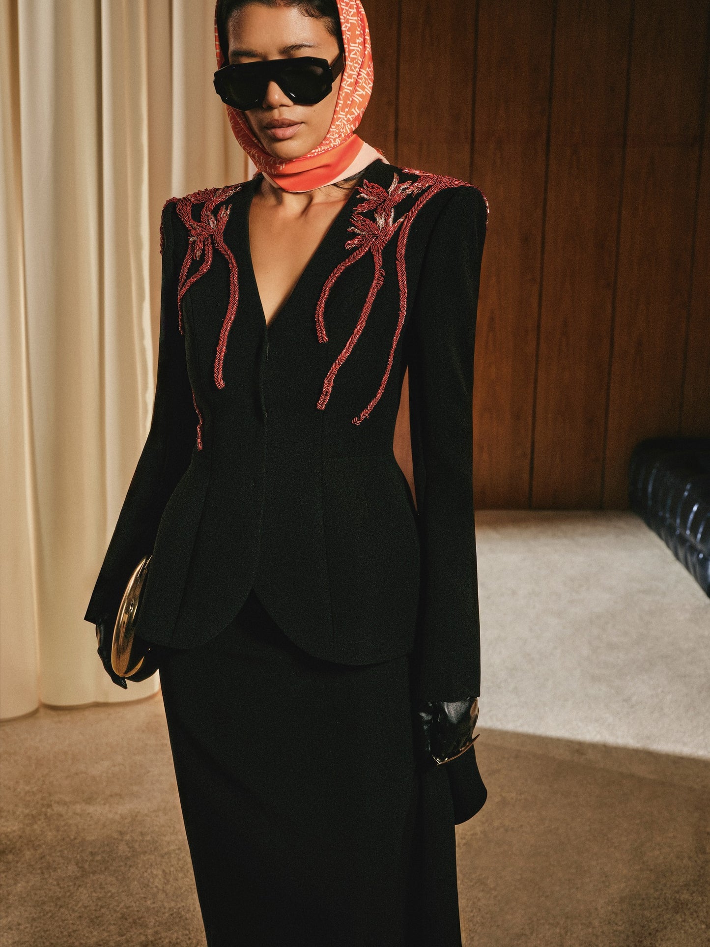 Person wearing a black outfit with red embroidery, sunglasses, and a headscarf in an indoor setting.